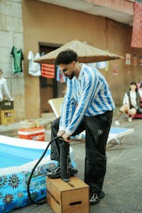Man in blue striped shirt inflating a pool outdoors with urban and casual setting.