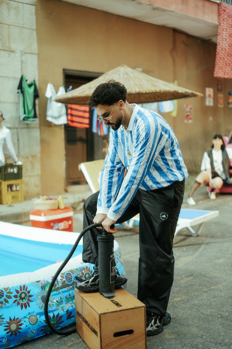 Man in blue striped shirt inflating a pool outdoors with urban and casual setting.