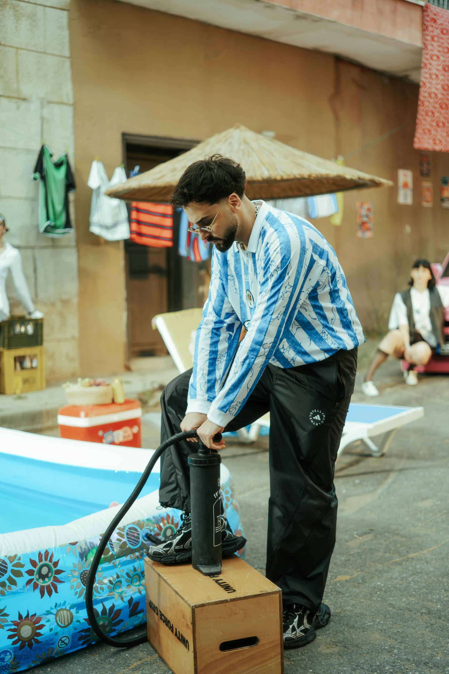 Man in blue striped shirt inflating a pool outdoors with urban and casual setting.